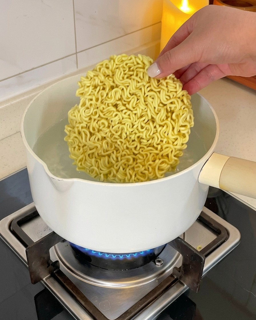 Una mano colocando un bloque circular de fideos de ramen instantáneo secos en una olla con agua hirviendo.