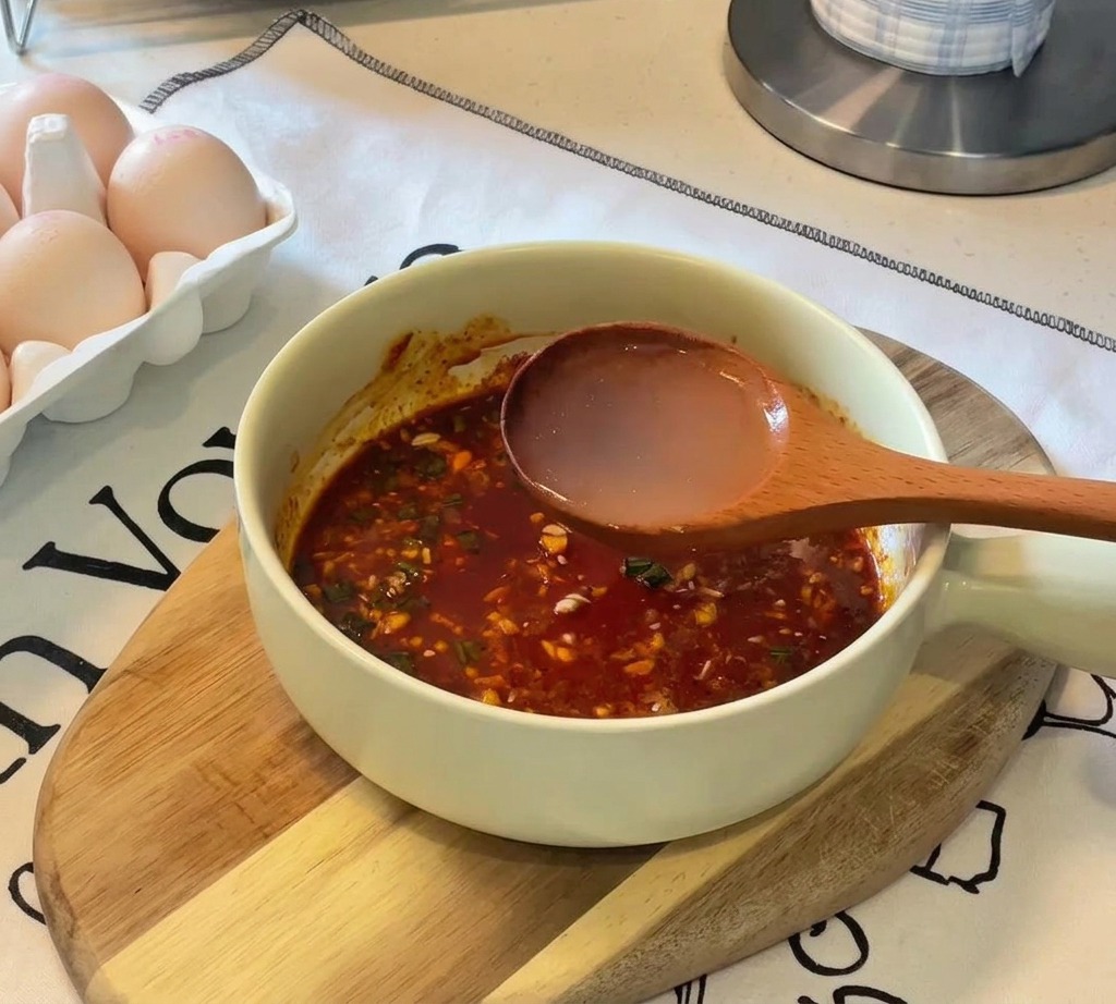 Un cucharón de madera vertiendo agua caliente transparente en un tazón con la pasta sazonadora de color rojizo.