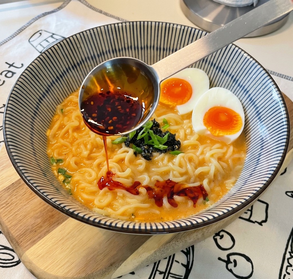 Una cuchara medidora metálica vertiendo aceite de chile de color rojo brillante con hojuelas visibles en un tazón de ramen.