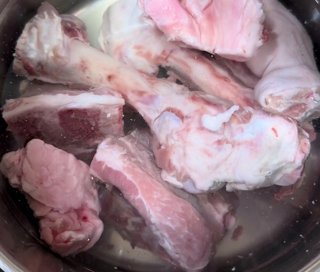 Huesos y manitas de cerdo crudos sumergiéndose en una olla grande de acero inoxidable llena de agua fría.
