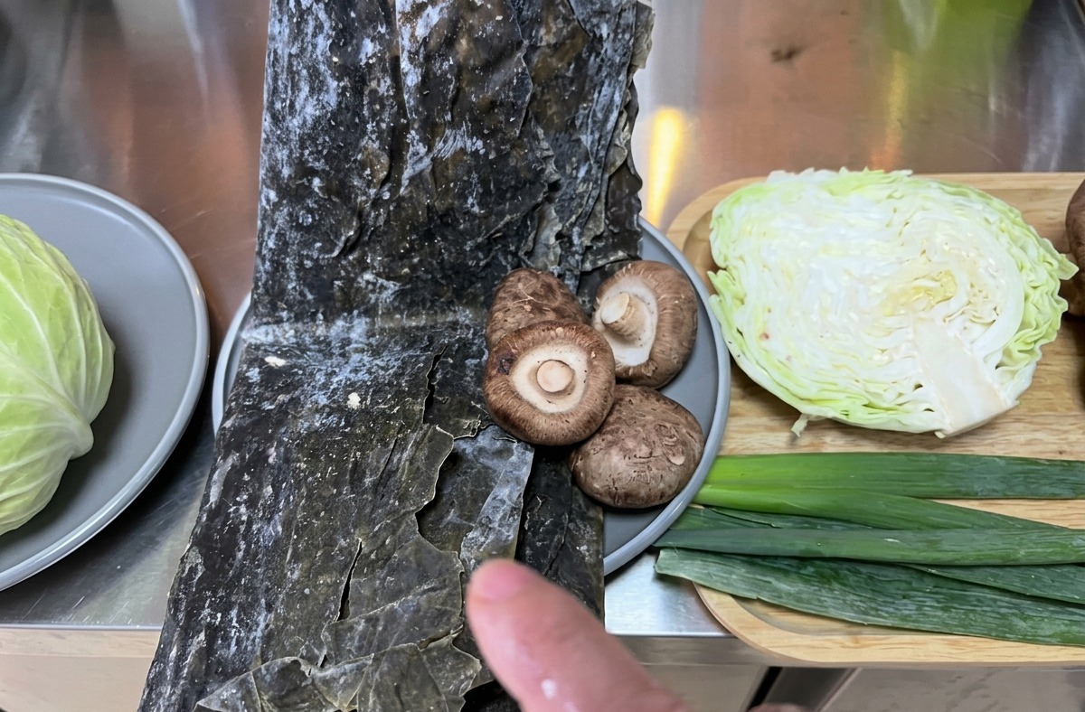 Ingredientes frescos que incluyen repollo, alga kombu y setas shiitake organizados en la encimera de la cocina.