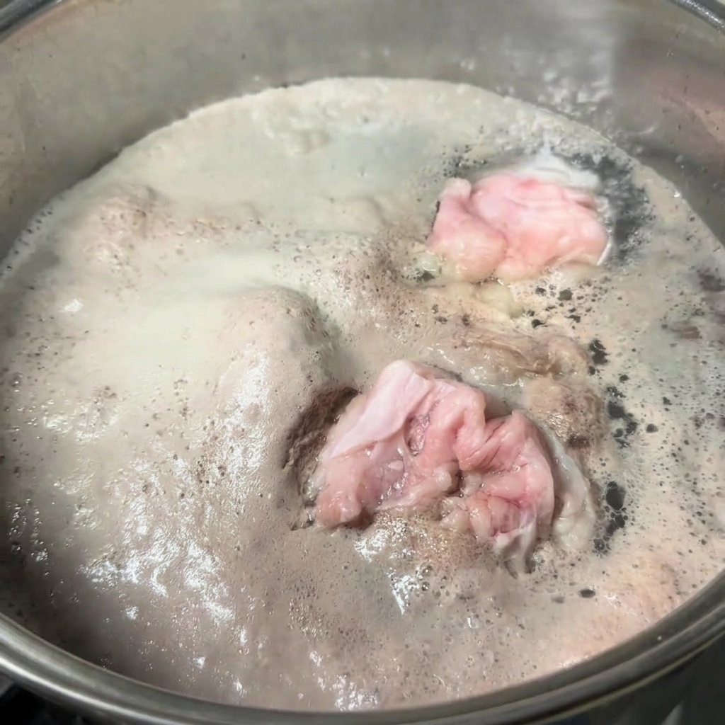 Una olla de acero inoxidable con agua hirviendo y huesos de cerdo cubierta por una capa gruesa de espuma gris.