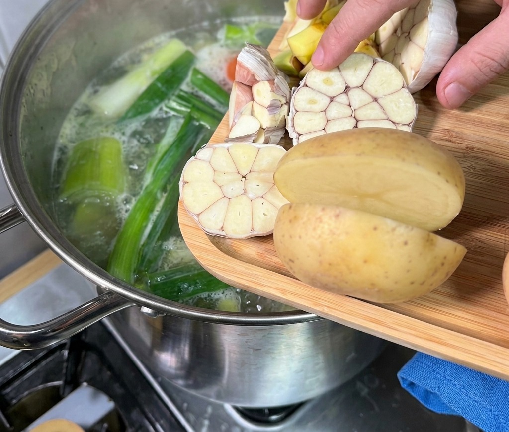Una mano agregar cabezas de ajo cortadas por la mitad y patatas desde una tabla de madera a una olla humeante de caldo.