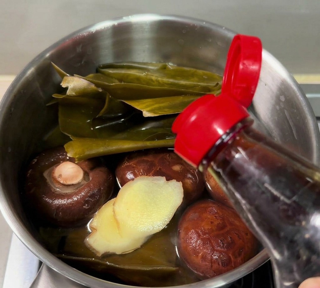 Salsa de pescado vertiéndose desde una botella en un cazo pequeño con kombu, setas shiitake y jengibre en rodajas.