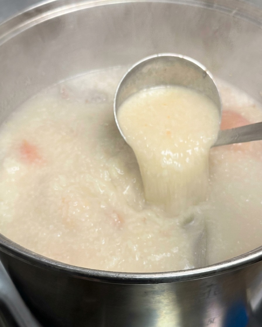 Un cucharón metálico levantando una porción de caldo tonkotsu espeso, opaco y de color blanco lechoso de una olla grande.