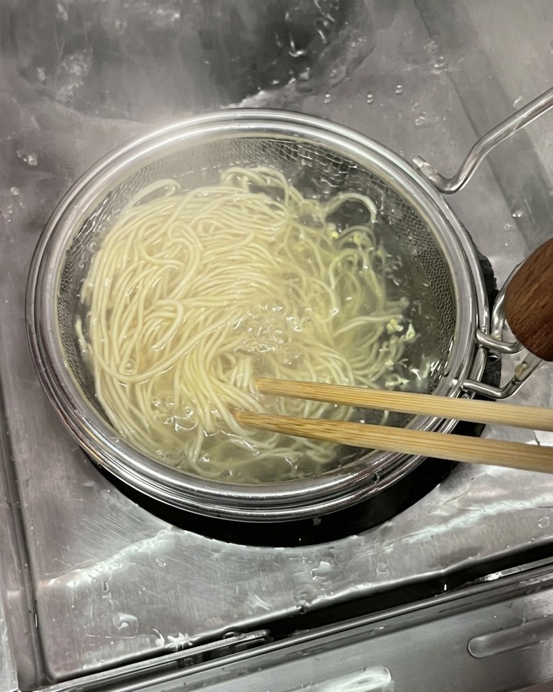 Un manojo de fideos finos y pálidos de trigo siendo levantados del agua hirviendo dentro de un colador de malla cilíndrico con palillos de madera.