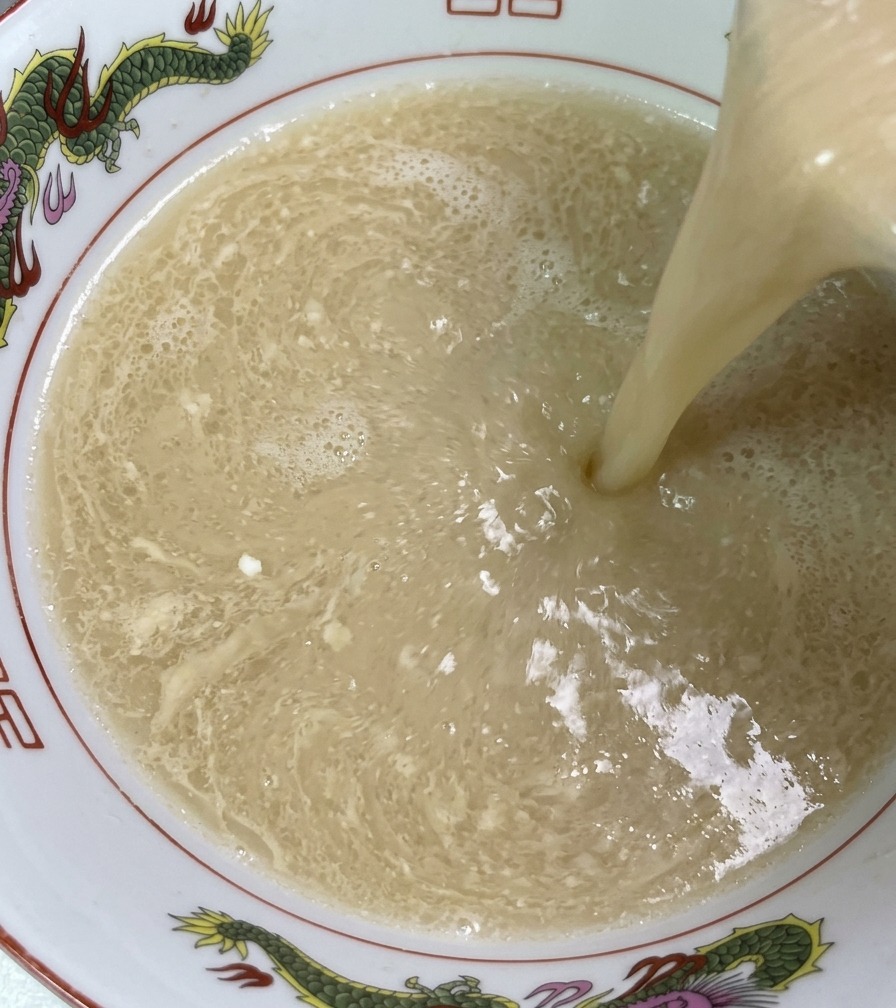Caldo humeante de huesos de cerdo de color marfil vertiéndose desde un cucharón en un tazón de ramen decorado.