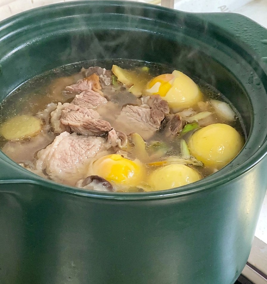 Una olla de barro verde con una sopa de huesos de cerdo burbujeante con trozos visibles de maíz y ñame hirviendo en la estufa.