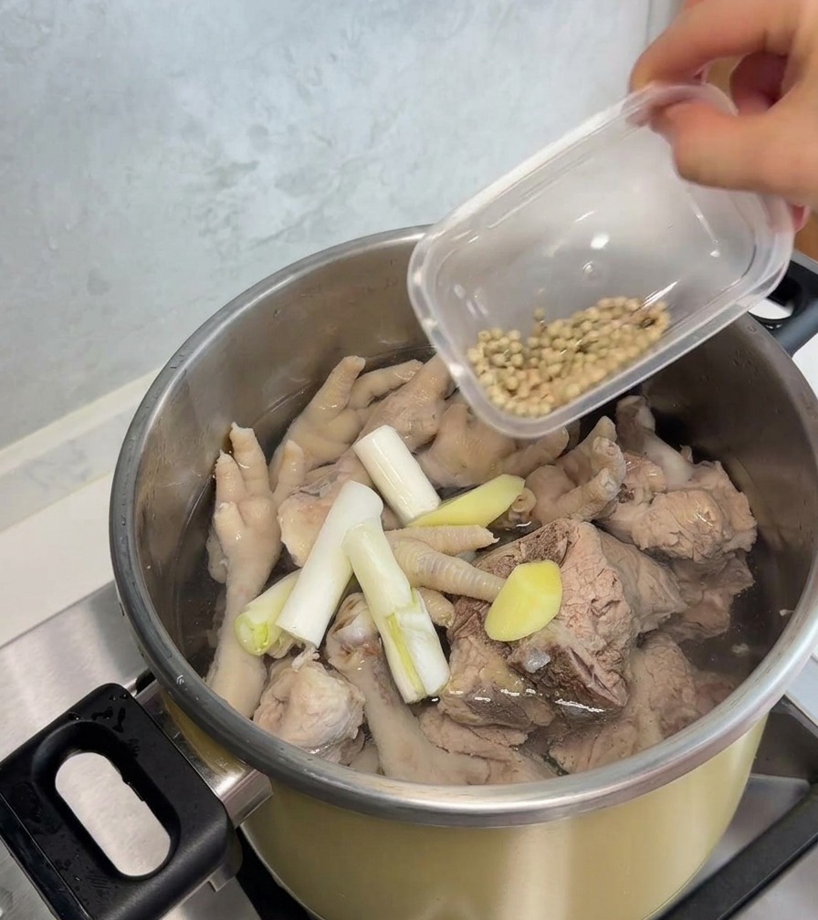 Una mano vertiendo granos de pimienta blanca sobre una olla de acero inoxidable que contiene patas de pollo, huesos de cerdo, cebollín y rodajas de jengibre.