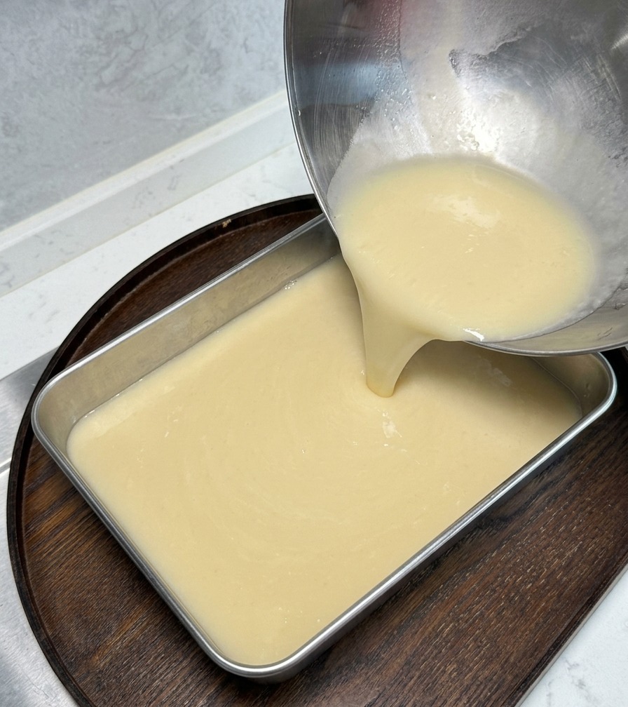 Caldo espeso de color crema vertiéndose desde un bol de metal hacia una bandeja rectangular.