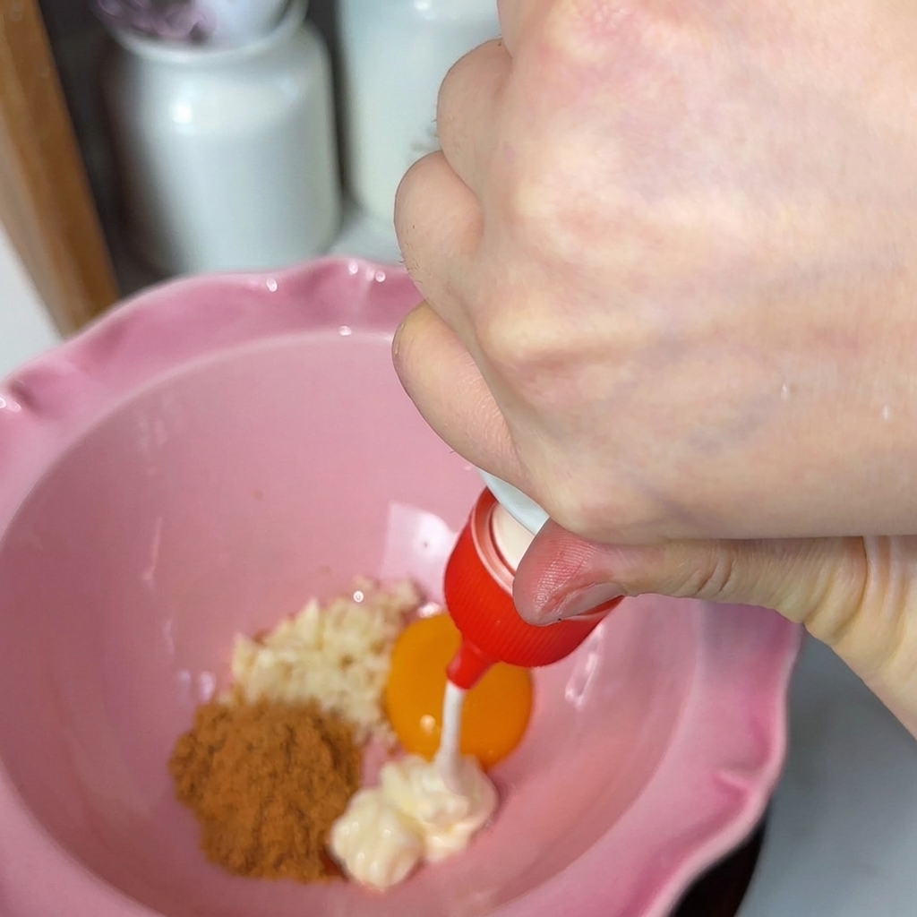 Mayonesa siendo añadida a un bol rosado que contiene ajo picado, una yema de huevo naranja brillante y polvo sazonador.
