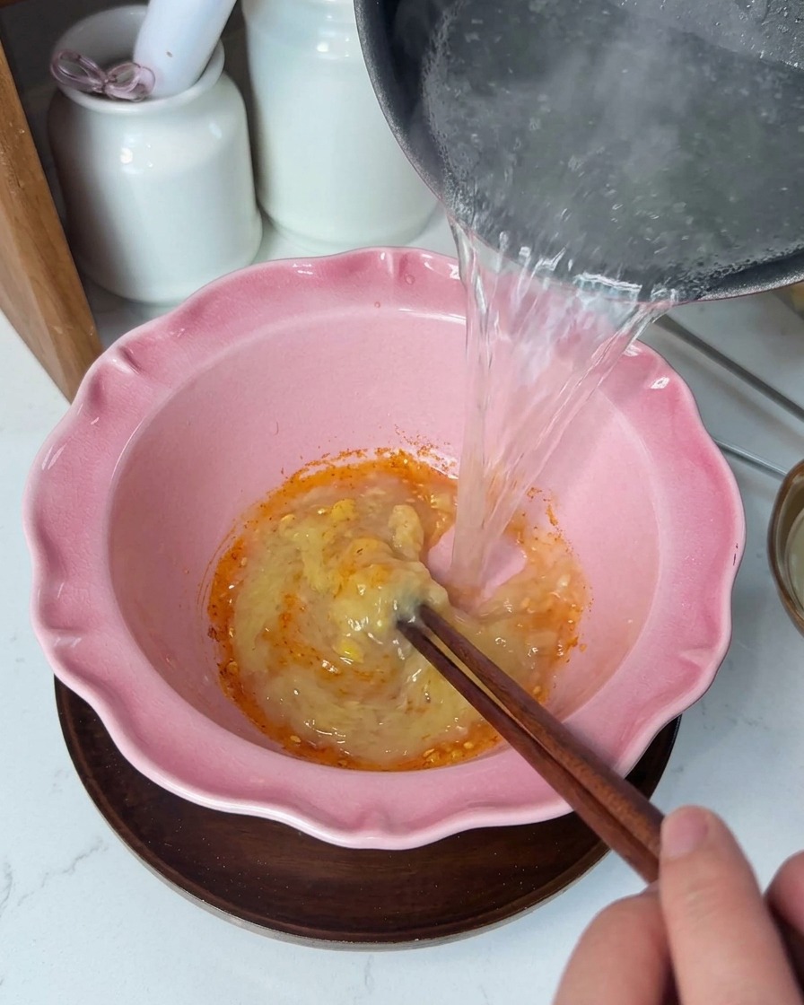 Líquido muy caliente cayendo en un bol rosado sobre una pasta amarilla y cremosa, mientras se remueve con palillos.