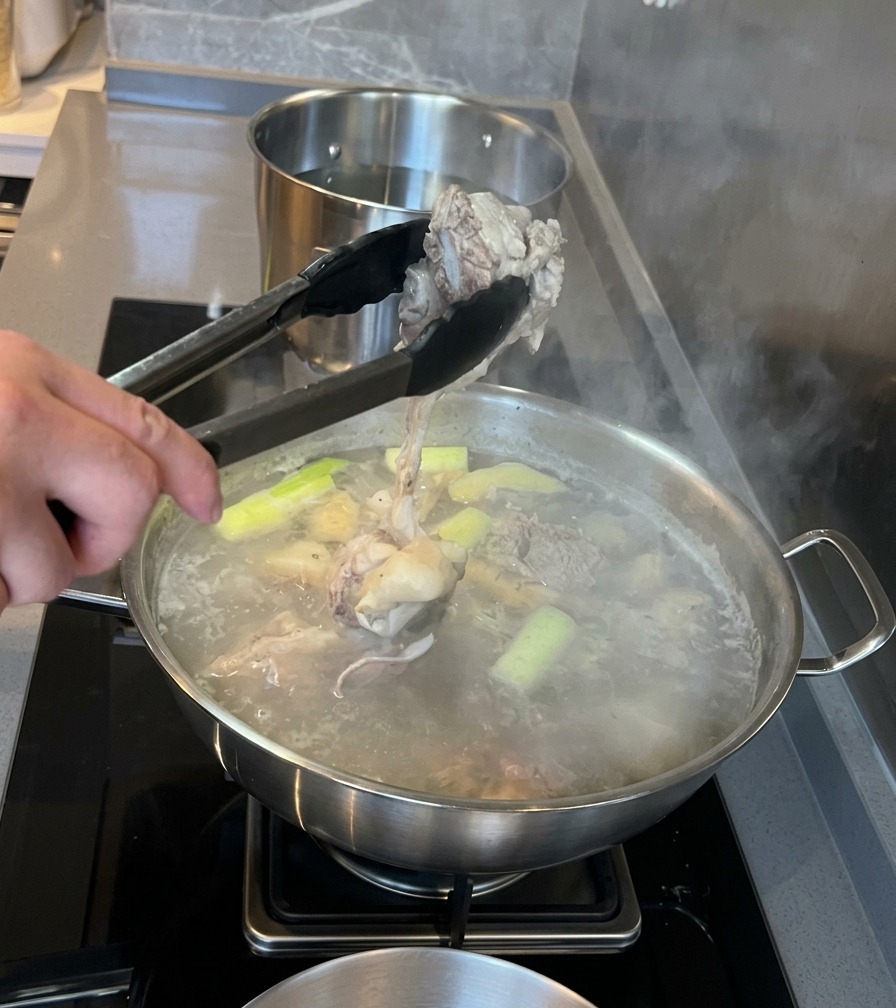 Unas pinzas de metal sacando un trozo de hueso de tuétano de cerdo blanqueado de una olla de agua hirviendo con hierbas aromáticas.