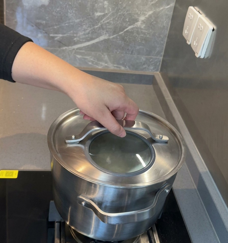 Una mano colocando una tapa de vidrio con borde de acero inoxidable en una olla grande para iniciar un largo proceso de cocción a fuego lento.