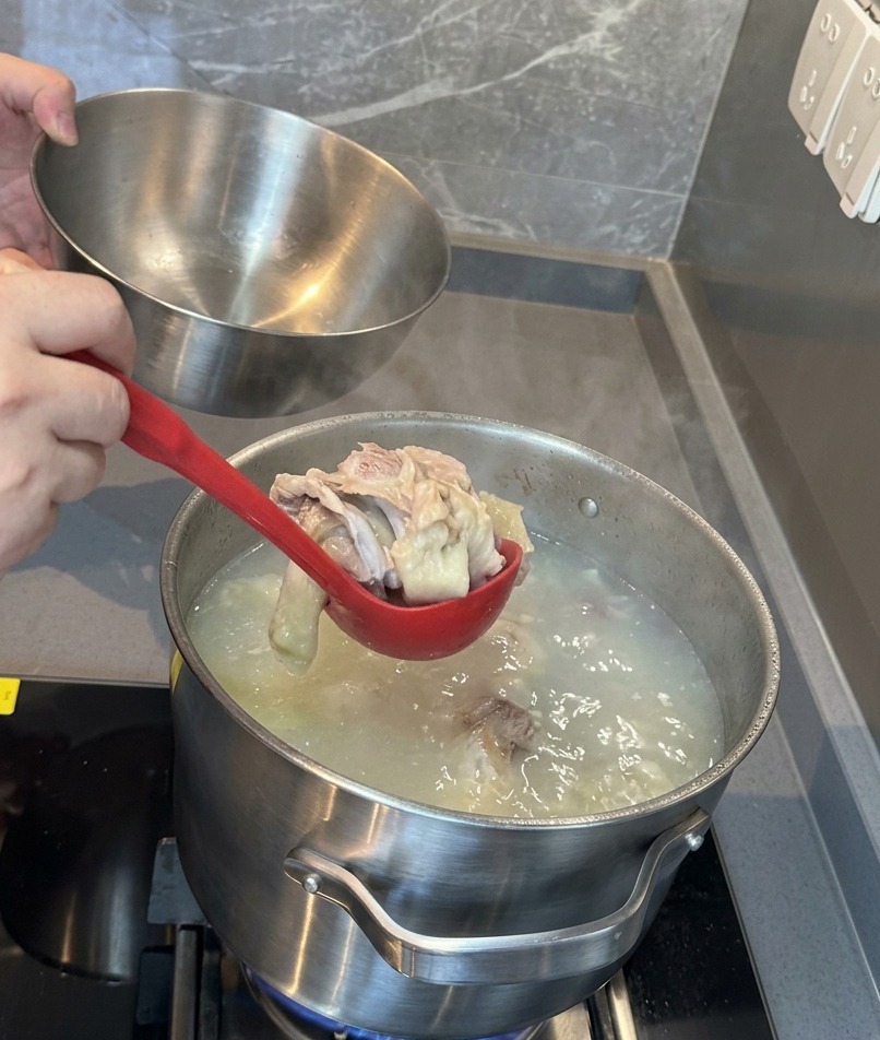 Un cucharón rojo sacando los huesos de cerdo y la carne ya ablandados de un caldo blanco lechoso para pasarlos a un bol de acero inoxidable.