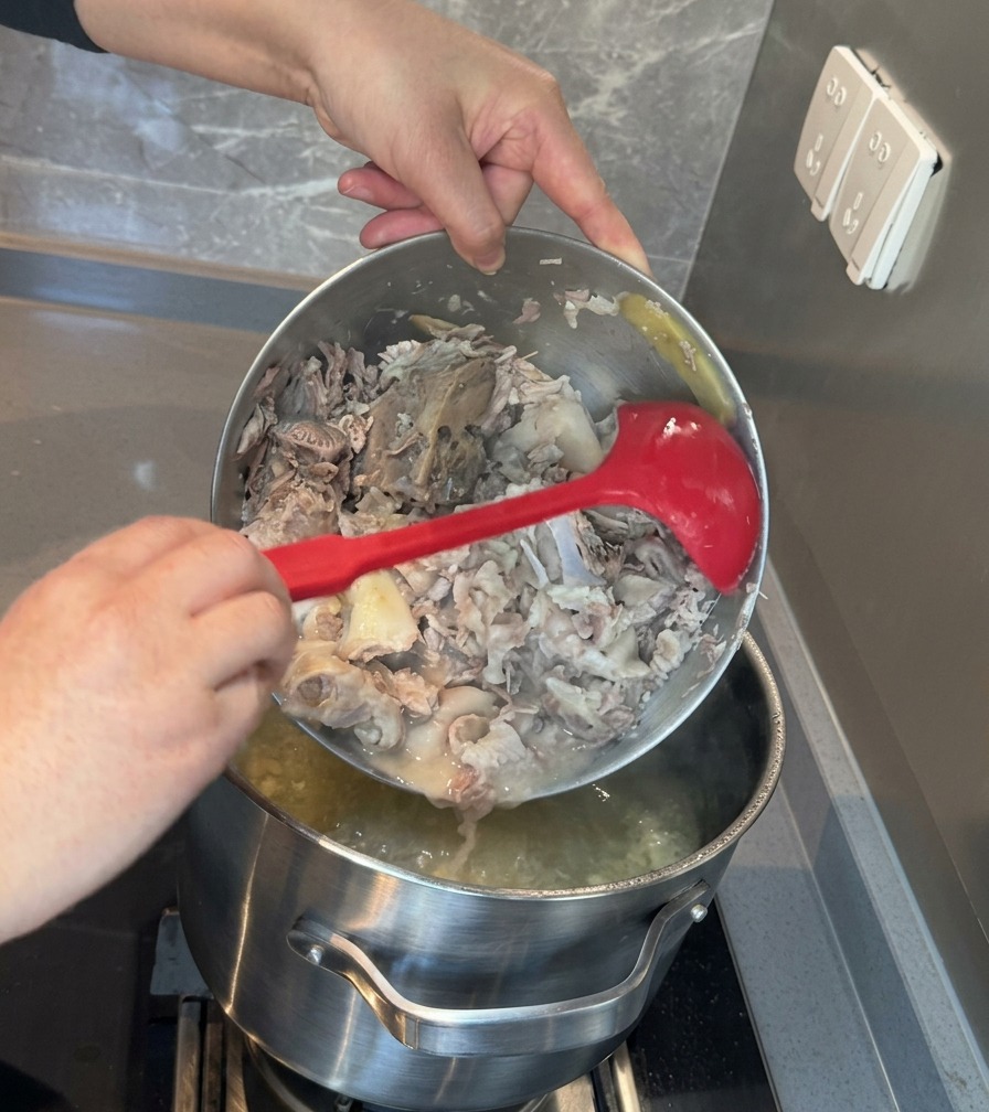El contenido de un tazón de metal, consistente en carne y huesos de cerdo machacados, se vierte de nuevo en una olla grande con caldo blanco.