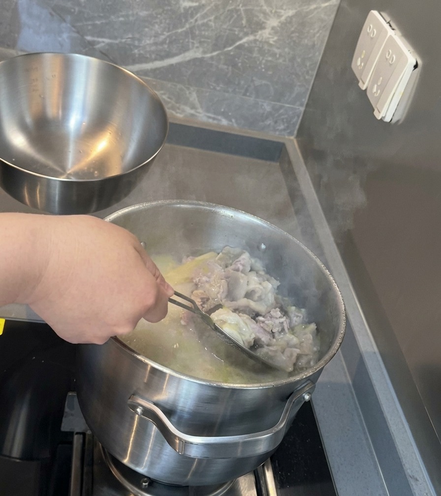 Un colador de malla de acero inoxidable filtrando trocitos de hueso y carne de una olla profunda con sopa de cerdo blanca y lechosa.