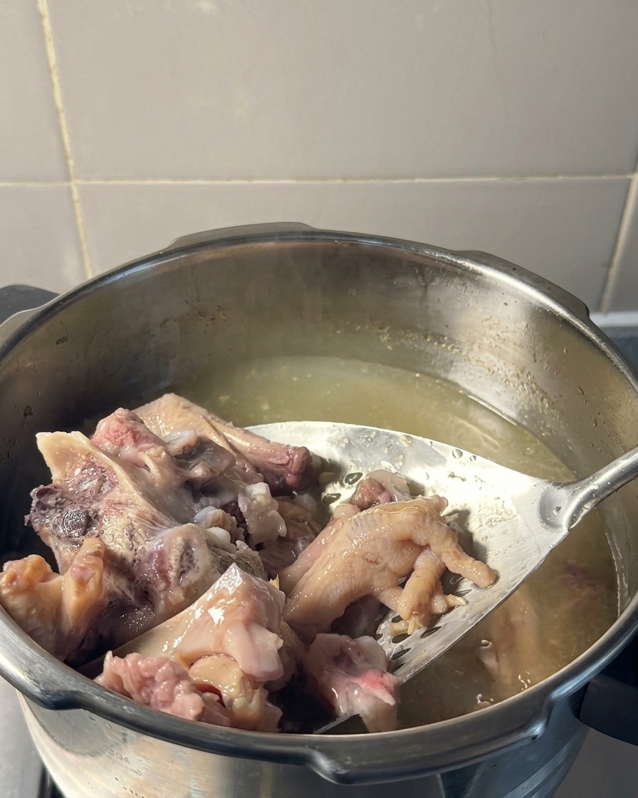 Una espátula de metal ranurada levantando carne de cerdo tierna y patas de pollo cocidas de una olla con caldo amarillo pálido.