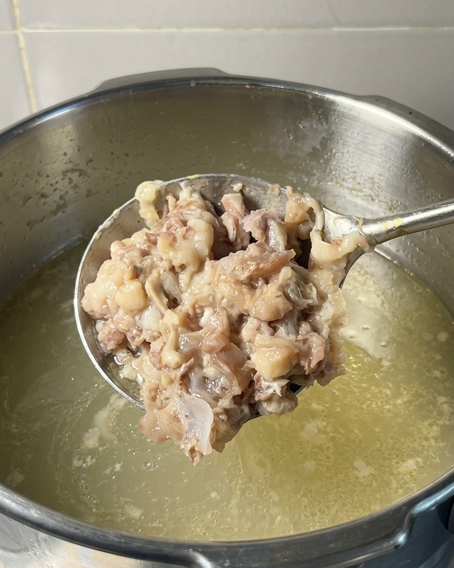 Un cucharón lleno de una pasta espesa de carne de cerdo y pollo machacada sostenido sobre una olla de caldo hirviendo.