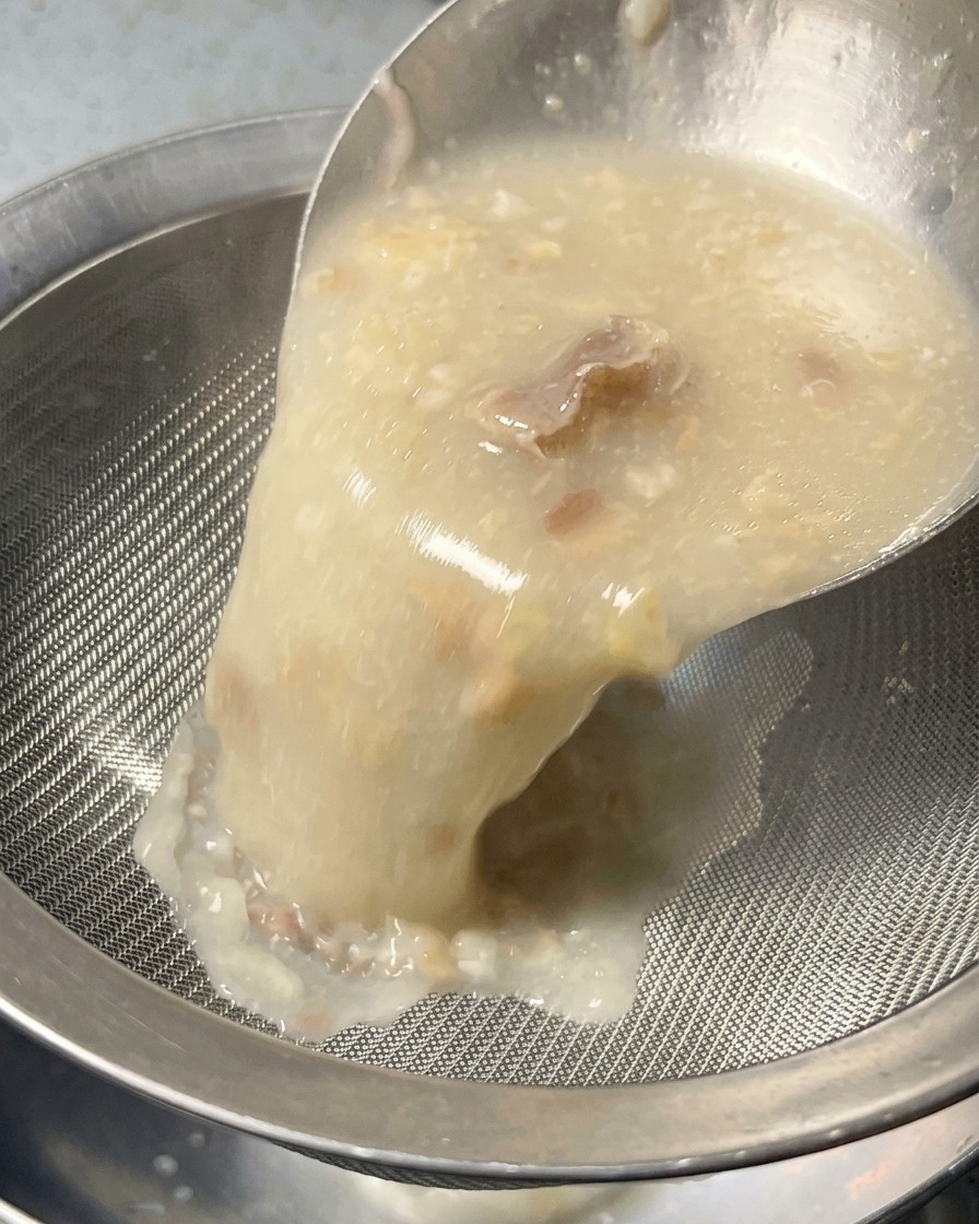 Un caldo blanco, espeso y opaco que se vierte a través de un colador de malla plateada en un bol, filtrando los trozos sólidos.