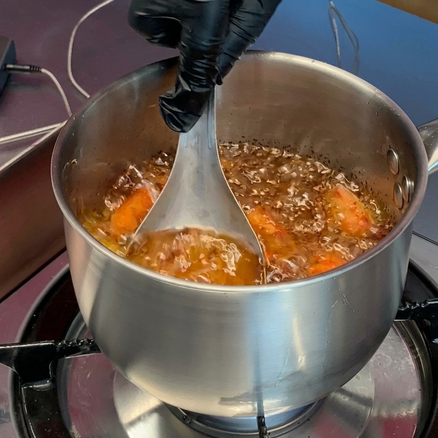 Una mano con guante presionando cabezas de camarón cocidas de color naranja con un cucharón de metal dentro de una olla con aceite caliente burbujeante.