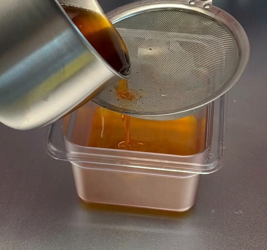 Vertiendo aceite de camarón de tono rojo anaranjado desde una olla de acero inoxidable a través de un colador de malla fina hacia un recipiente rectangular transparente.