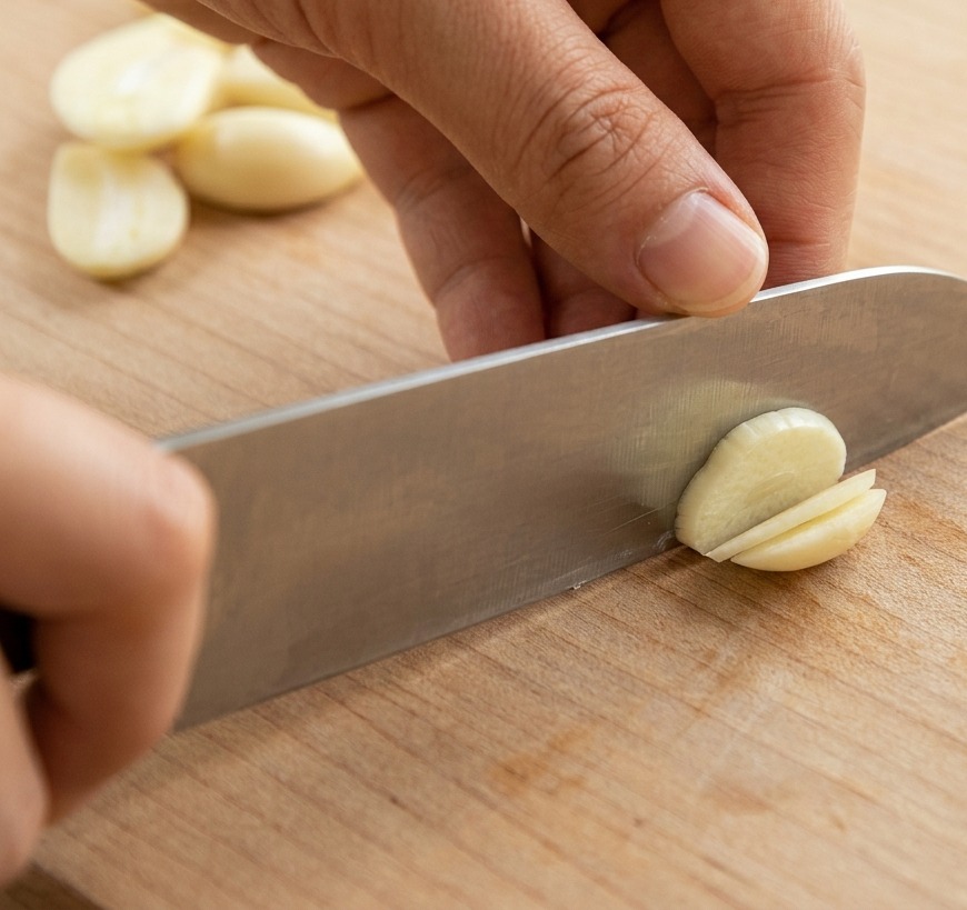 Un plano de cerca de unas manos usando un cuchillo afilado para cortar dientes de ajo enteros en rodajas finas sobre una tabla de cortar de madera.