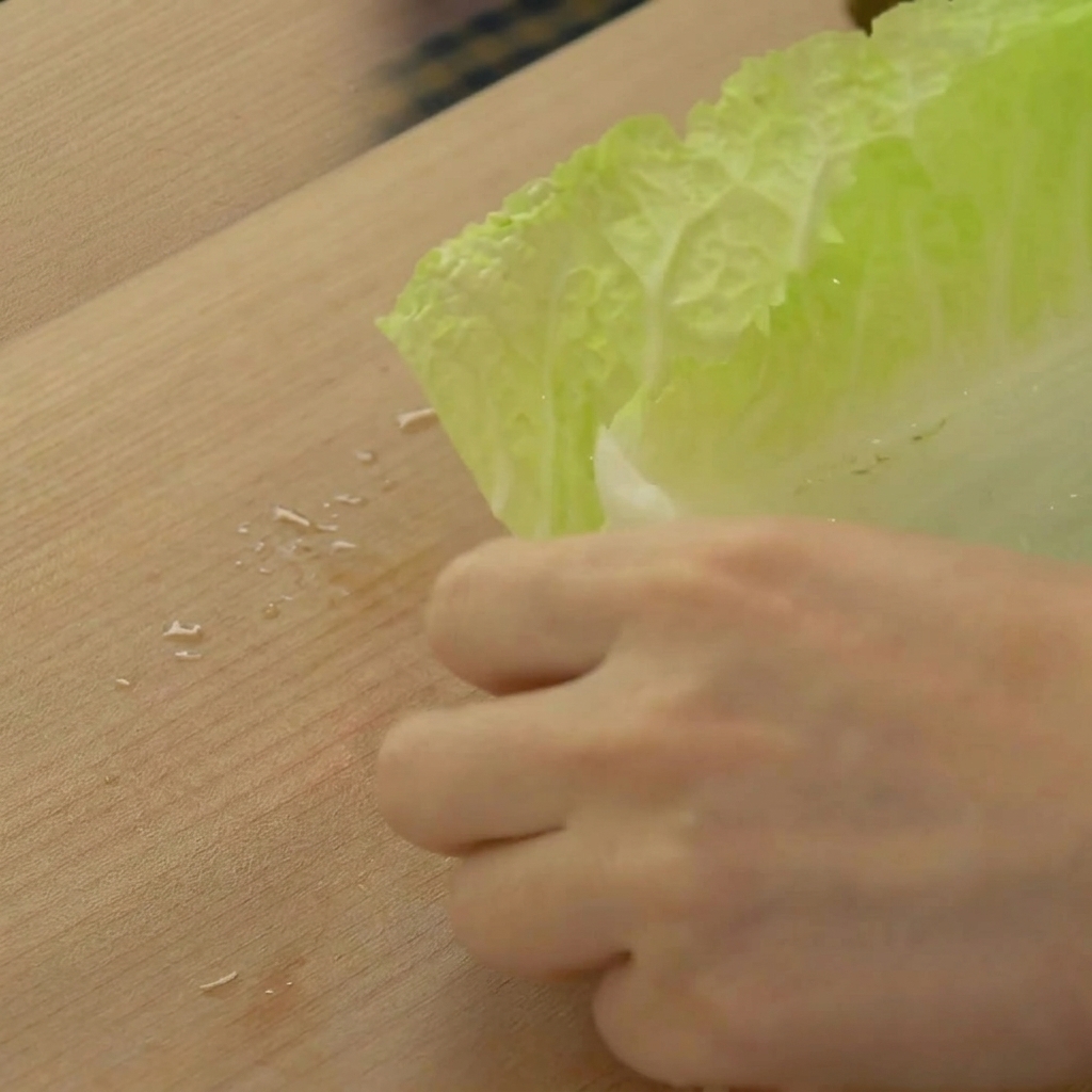 Manos cortando un fajo de hojas de col china de color verde claro para hacer tiras anchas sobre una tabla de cortar de madera.