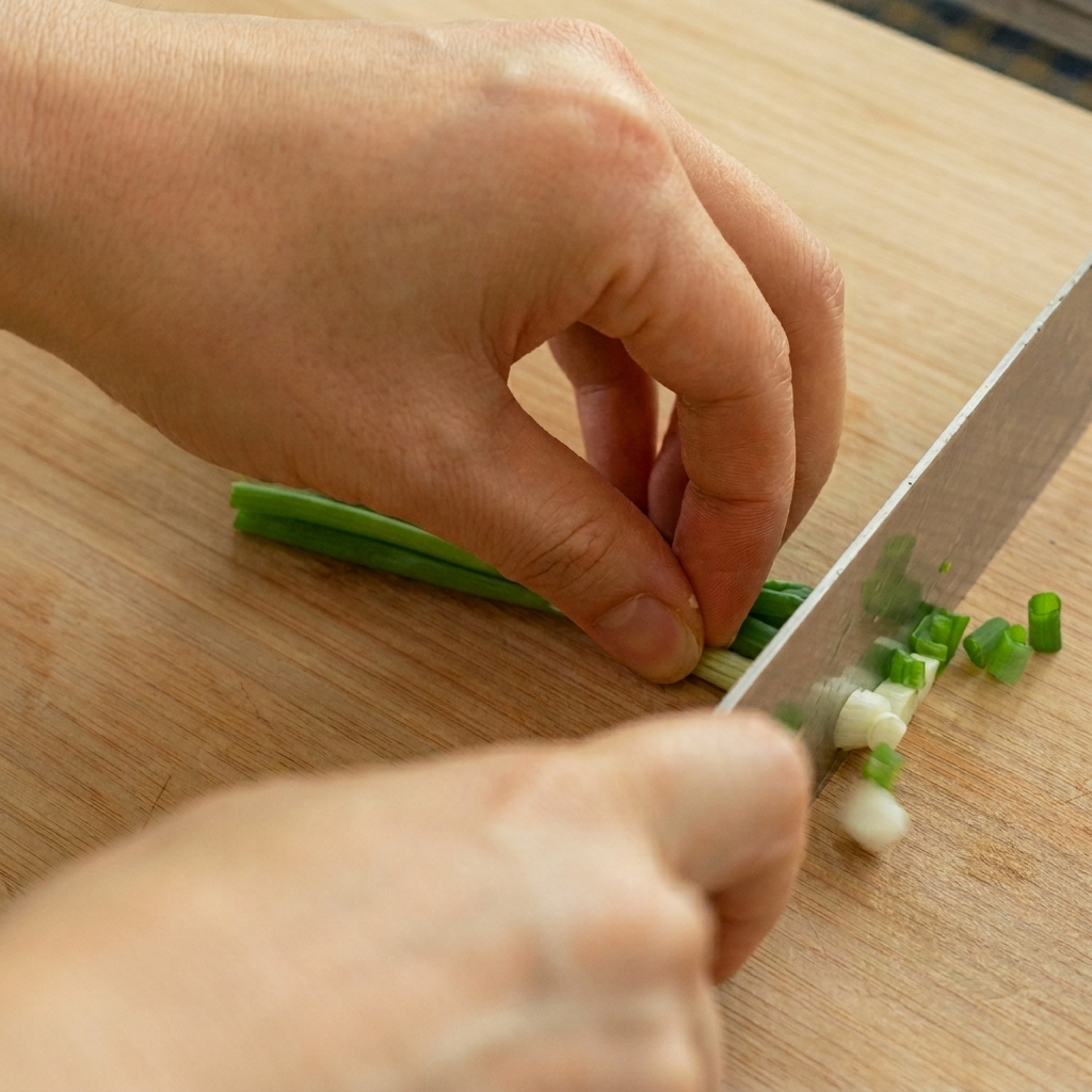 Un plano de cerca del cuchillo de un chef picando finamente un manojo de tallos de cebolleta en pequeños aros verdes y blancos.