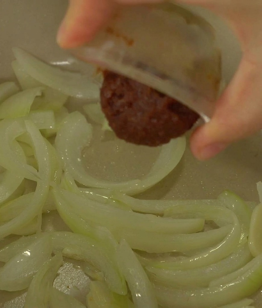 Una mano vertiendo una cucharada de pasta de soja fermentada de color marrón oscuro desde un cuenco pequeño hacia una olla con cebollas sofritas translúcidas.