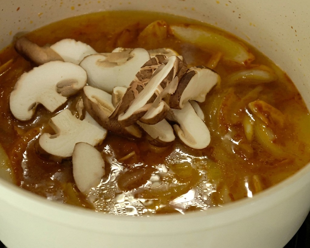 Setas recién cortadas y trozos de col reposando en la superficie de un rico caldo de ramen de color anaranjado que hierve a fuego lento en una olla blanca.