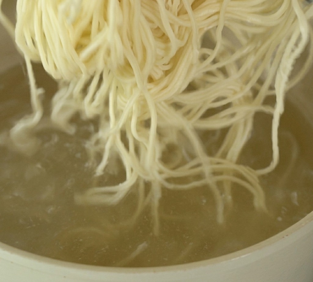 Un manojo de fideos ramen delgados y claros siendo sumergidos en una olla de agua hirviendo a borbotones.