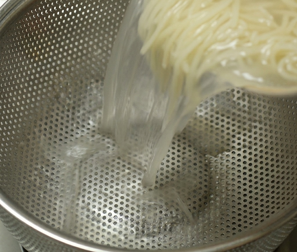 Una olla con fideos ramen cocidos vertiéndose en un colador de acero inoxidable para escurrir el líquido de cocción caliente.