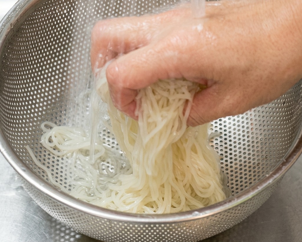 Una mano sacudiendo fideos ramen blancos en un colador de acero inoxidable bajo un chorro de agua fría en un fregadero.