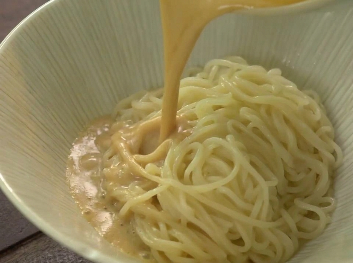 Un chorro de caldo de ramen caliente y beige cremoso vertiéndose sobre una montaña de fideos en un tazón de cerámica de color claro.
