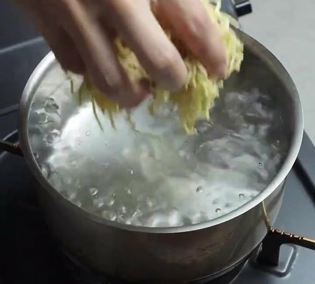 Una mano soltando un nido de fideos ramen frescos y amarillos en una olla de acero inoxidable con agua hirviendo a borbotones.