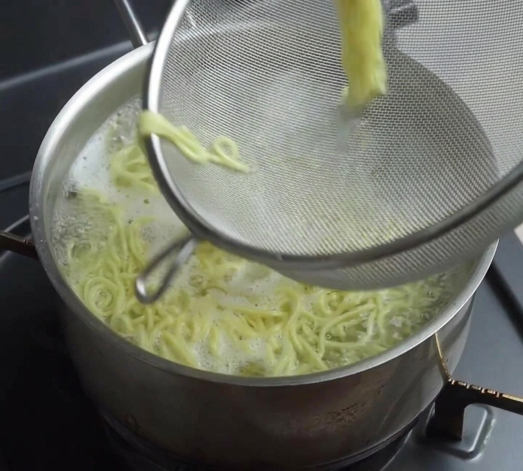 Un colador de malla de alambre levantando una porción de fideos ramen amarillos cocidos de una olla humeante.