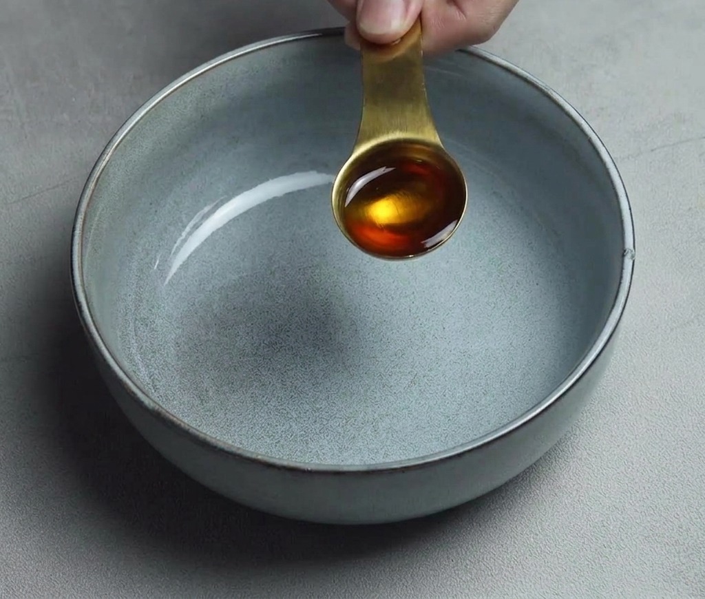 Una cucharada dorada de aceite de sésamo vertiéndose en el fondo de un tazón de cerámica gris limpio y vacío.