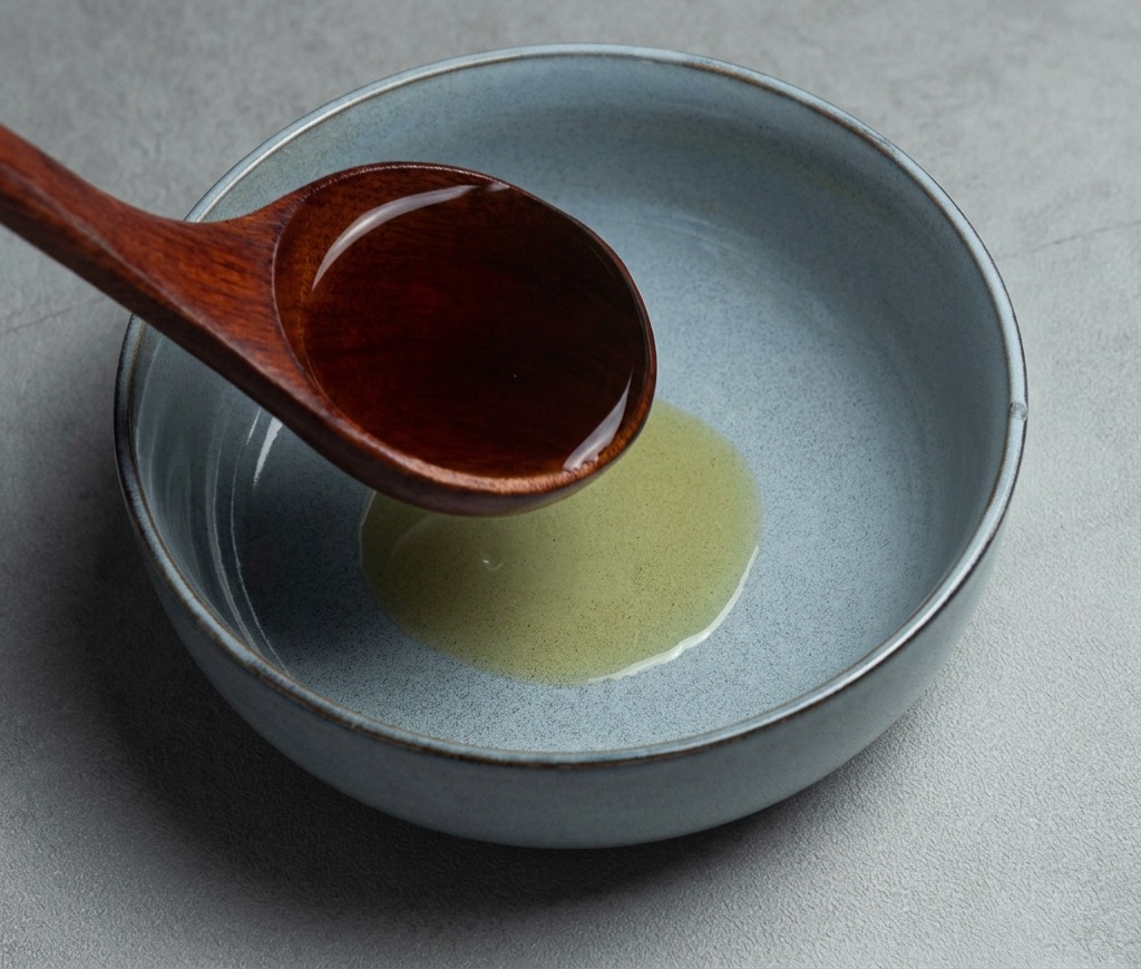 Un cucharón de madera oscura vertiendo caldo caliente de shoyu ramen en un tazón de cerámica gris que contiene aceite de sésamo.