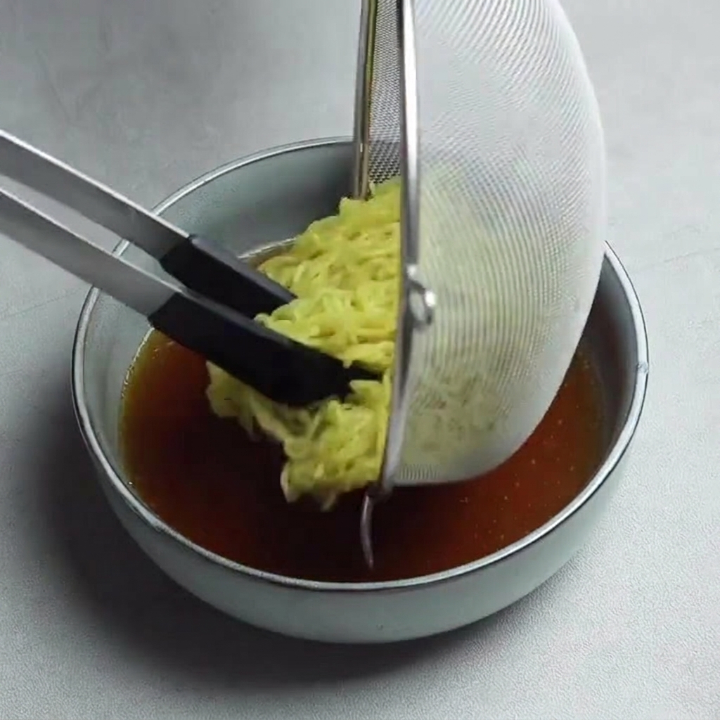 Fideos ramen amarillos siendo colocados desde un colador de malla en un tazón de caldo oscuro y sabroso usando pinzas de mango negro.