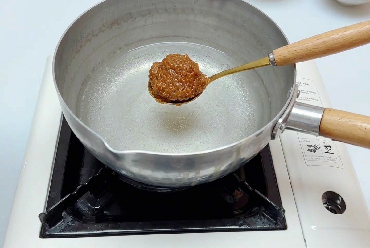Una cuchara dorada con una porción de la mezcla de miso introduciéndose en una olla de acero inoxidable con agua sobre la estufa.