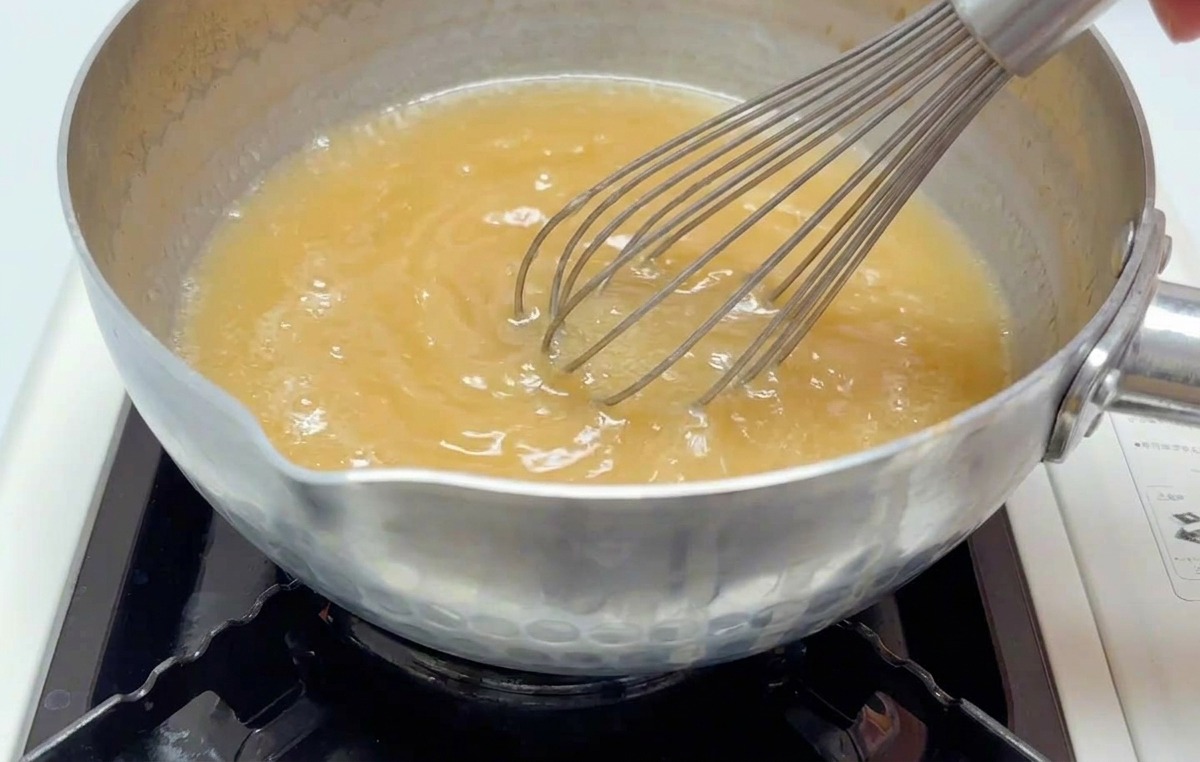 Una mano batiendo un caldo de miso de color marrón claro en una cacerola pequeña sobre un quemador encendido.