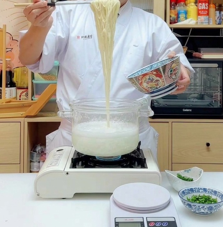 Una porción de fideos de ramen largos y amarillos sostenidos en alto con palillos al salir de una olla humeante.