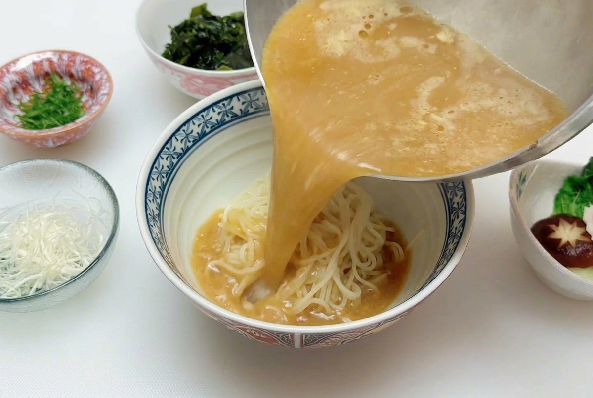 Un flujo continuo de caldo de miso caliente de color marrón dorado vertiéndose desde una cacerola sobre un nido de fideos de ramen en un tazón.