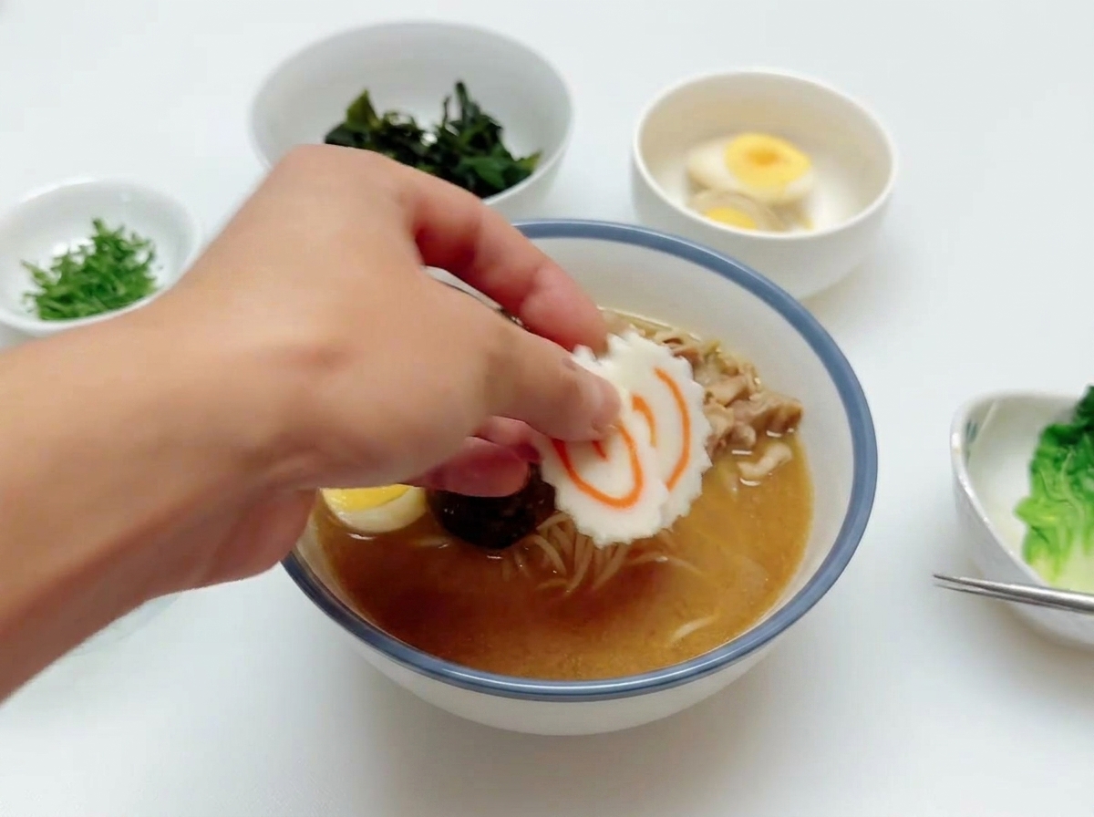 Una mano añadiendo una rebanada de pastel de pescado narutomaki con su espiral rosa sobre un tazón de ramen que ya tiene carne y huevo.