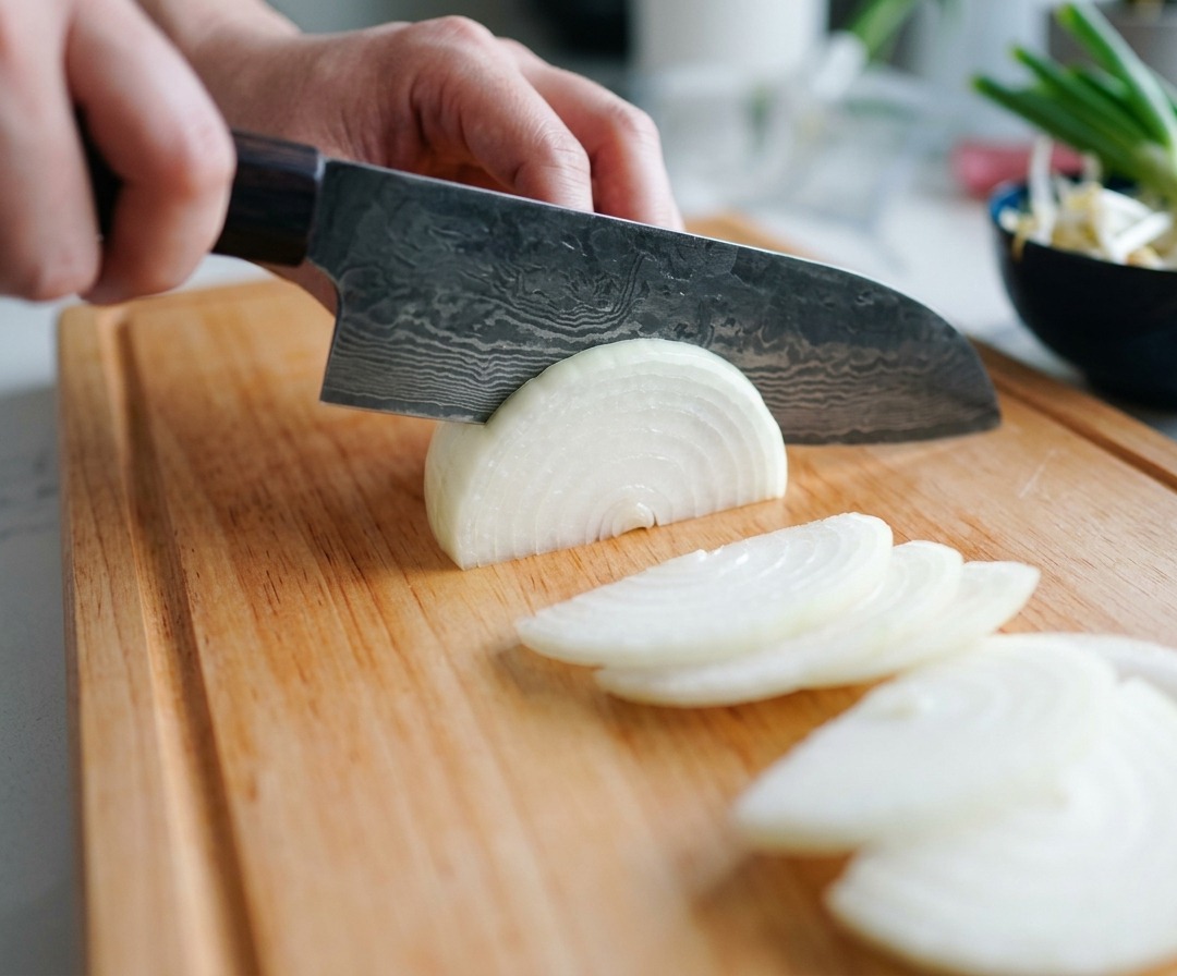 Una persona usando un cuchillo de chef de acero de Damasco para cortar una cebolla blanca en finas lunas sobre una tabla de madera.
