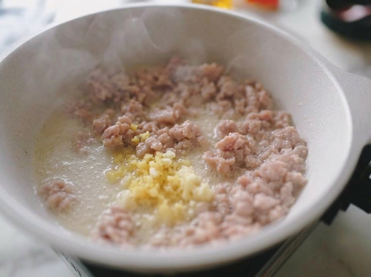 Ajo y jengibre picados mezclándose con la carne de cerdo picada y dorada en una sartén blanca, con vapor subiendo.