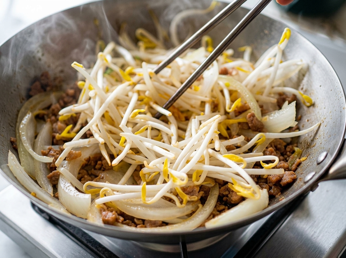 Un gran puñado de brotes de soja blancos que se echan en un wok con carne picada dorada y cebolla laminada.