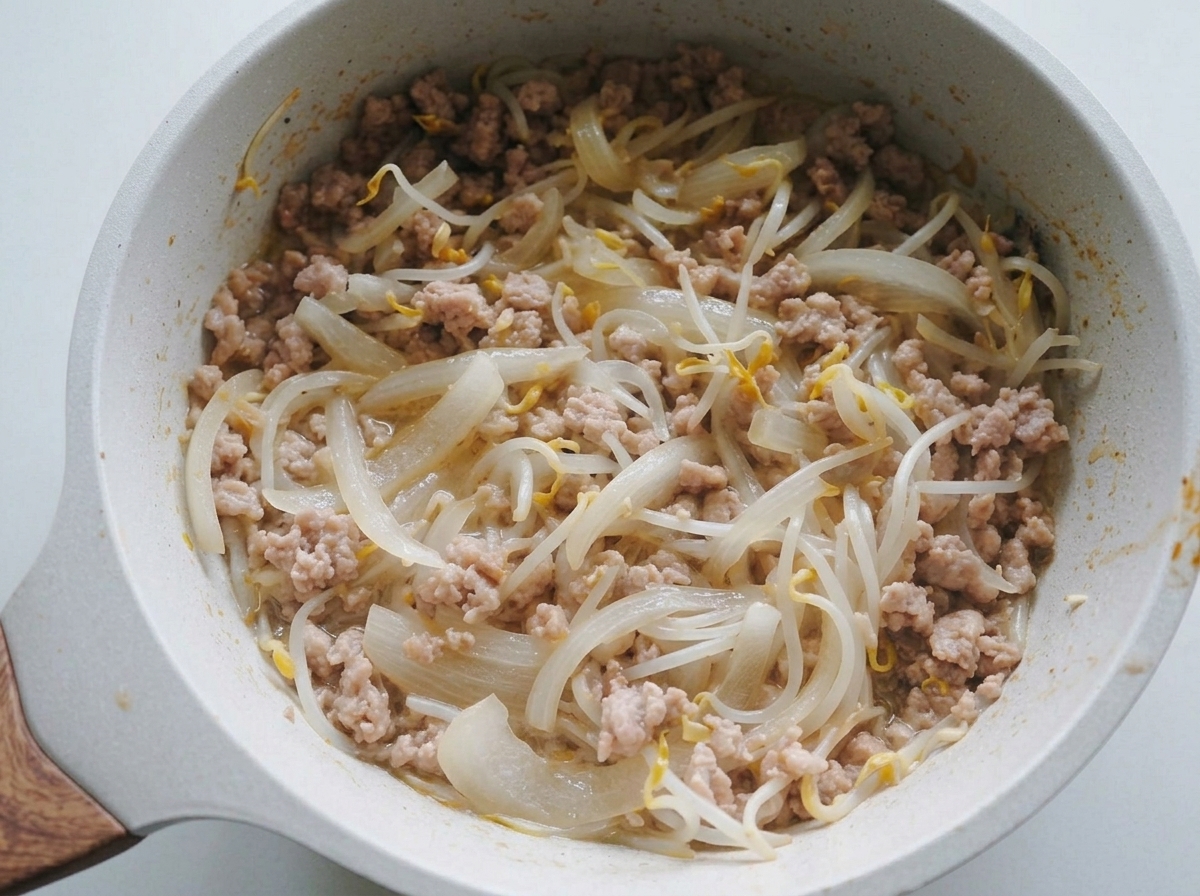 Una mezcla de cebolla pochada, brotes de soja y carne de cerdo picada removiéndose en una sartén de color claro.