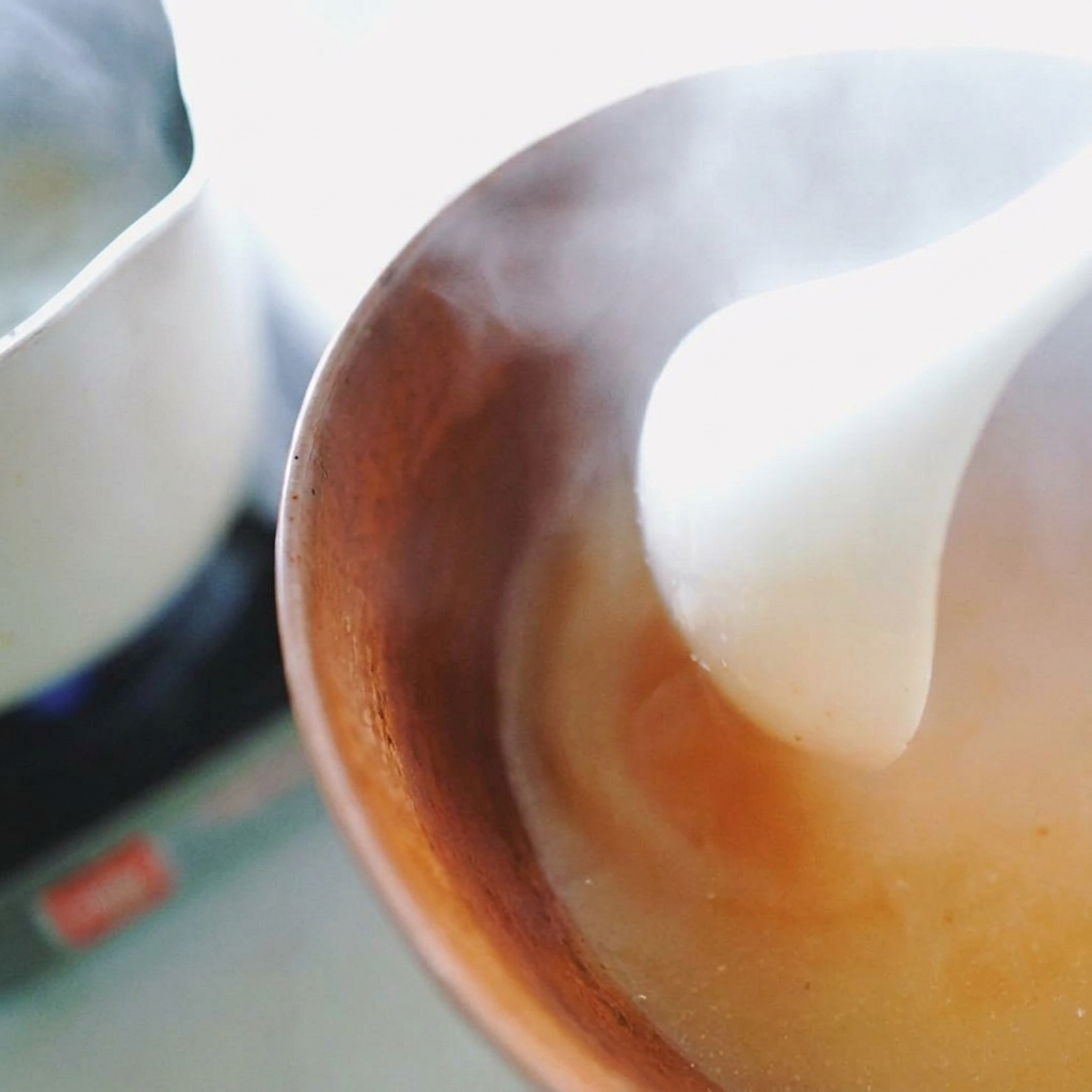 Un cucharón blanco vertiendo líquido caliente en un tazón marrón para disolver la pasta de miso en un caldo humeante.